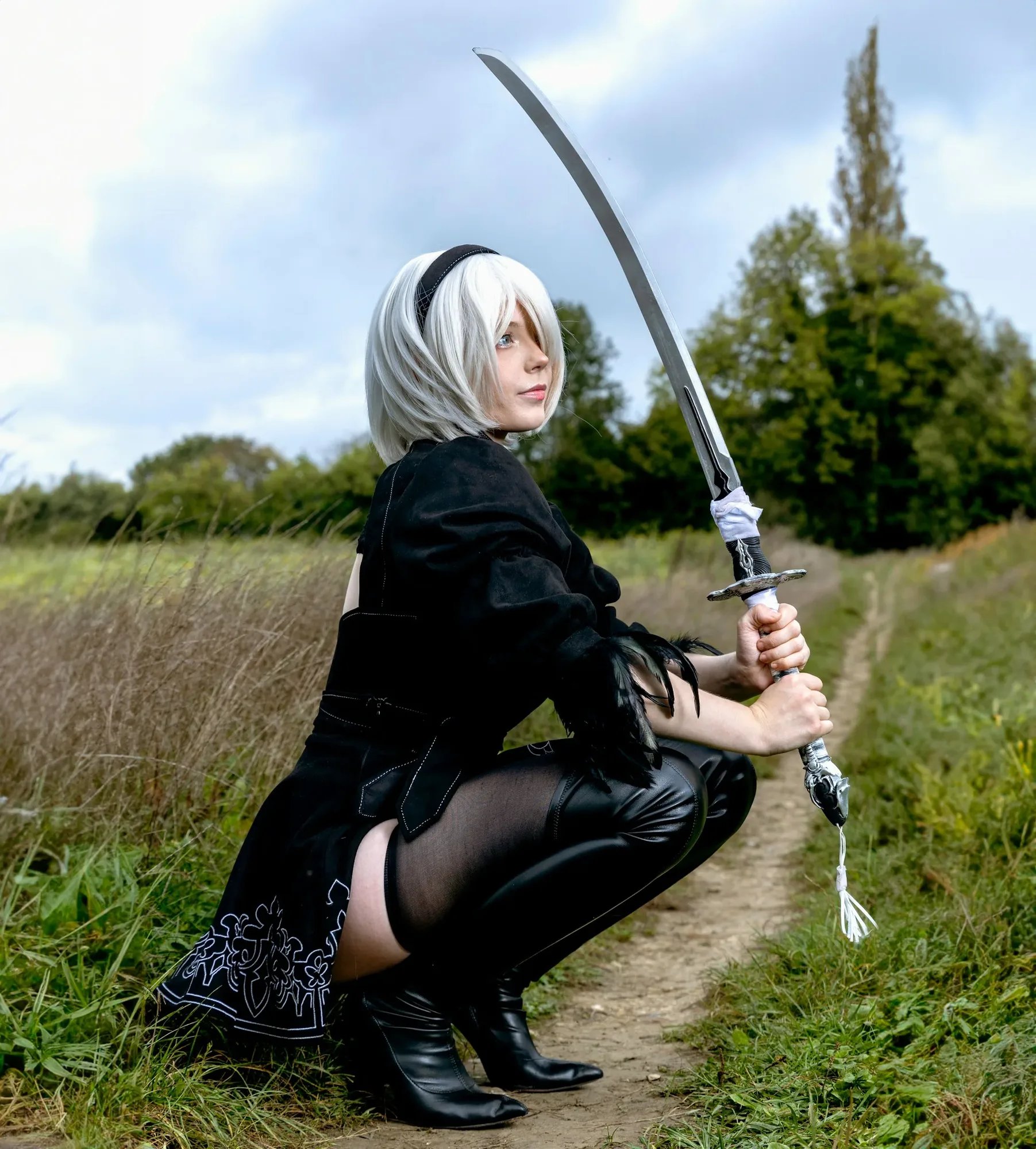 Cosplayer dressed in black kneeling outdoors with a sword.